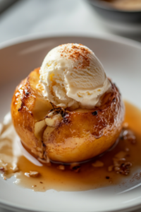 A close-up shot of a warm baked apple on a white plate over the white marble cooktop, topped with a scoop of melting vanilla ice cream. A spoon takes a bite, revealing the soft, spiced apple center paired with the crispy oat topping.
