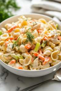 A serving bowl filled with freshly made crab pasta salad, garnished with sprigs of fresh dill. The bowl is placed on the white marble cooktop, with a spoon resting beside it, ready for serving.
