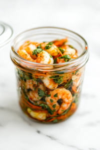 A chilled glass jar on the white marble cooktop, freshly taken from the fridge. A spoon lifts marinated shrimp from the jar, glistening with infused oil and seasonings, ready to be served.