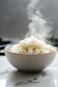 A close-up shot of a white bowl filled with warm, fluffy Calrose rice, placed on the white marble cooktop. Steam rises from the bowl, highlighting its perfect texture, ready to be served as a side dish or a base for flavorful meals.