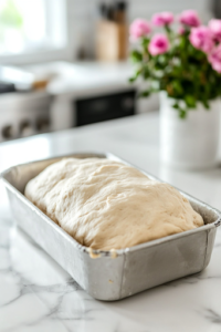 A close-up shot of the dough shaped into a smooth loaf and placed in a greased loaf pan on the white marble cooktop. The dough is evenly distributed, ready for its final rise before being baked to perfection.