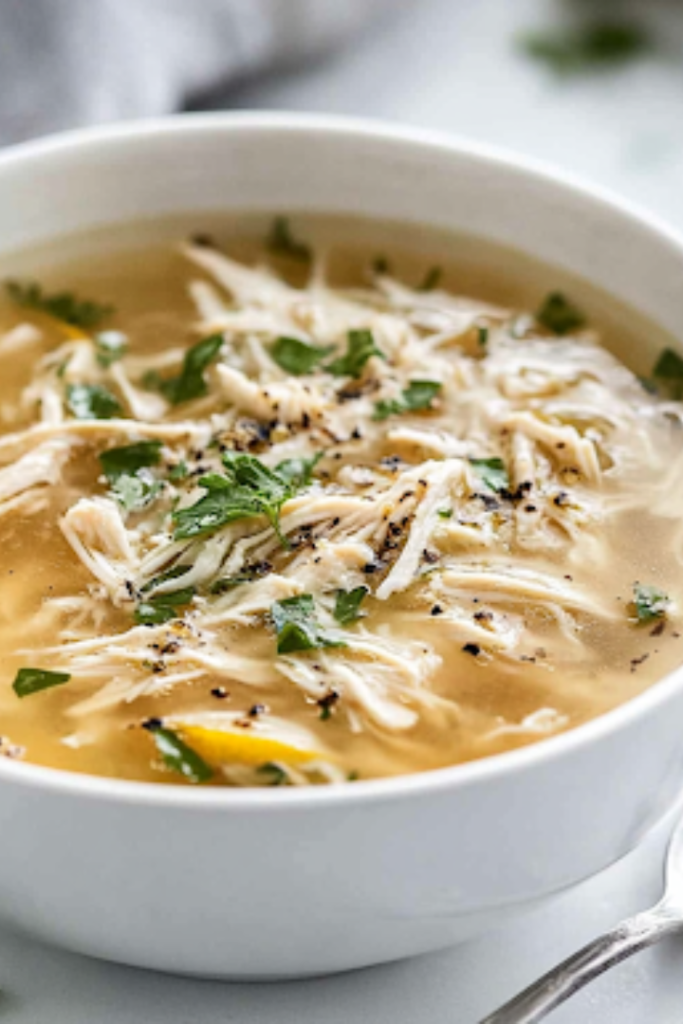A close-up shot of a steaming bowl of chicken soup with lemon and spices on a white marble cooktop. The dish is garnished with fresh herbs, adding a pop of color and aroma. A spoon rests beside the bowl, ready for a comforting meal