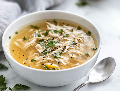 A close-up shot of a steaming bowl of chicken soup with lemon and spices on a white marble cooktop. The dish is garnished with fresh herbs, adding a pop of color and aroma. A spoon rests beside the bowl, ready for a comforting meal