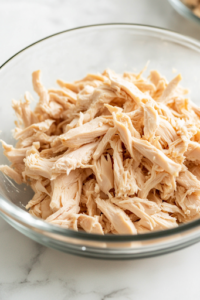 A close-up shot of a glass bowl on a white marble cooktop, filled with shredded cooked chicken breasts. A fork pulls apart the tender pieces, revealing their juicy texture and delicate fibers.