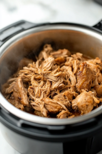 A black Instant Pot on a white marble cooktop, now open. A fork shreds the tender, juicy chicken thighs on a wooden cutting board before being returned to the pot. The steaming rice mixture absorbs all the flavors.