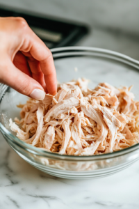 A glass mixing bowl sits on a white marble cooktop, filled with tender, shredded rotisserie chicken. A hand gently separates the meat into bite-sized pieces, preparing it for the salad.