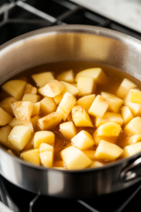 A close-up shot of a saucepan on a stovetop, filled with diced Granny Smith apples, sugar, cornstarch, lemon juice, and cinnamon simmering in water. The mixture appears glossy and thickened, with the apples softening into a rich filling.