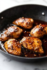 Chicken breasts simmer in a thickened maple glaze in a black skillet on a white marble cooktop. A spoon drizzles the glossy, golden sauce over the chicken, ensuring every piece is well coated.