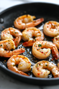 A black skillet on a white marble cooktop with large shrimp sizzling in olive oil and red pepper flakes. The shrimp are turning pink and slightly curled as they cook, releasing a fragrant, spicy aroma.