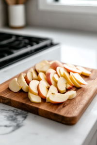 A wooden cutting board on the white marble cooktop with peeled and thinly sliced apples neatly arranged. A hand is tossing the apple slices in a glass mixing bowl with granulated sugar, lemon juice, and cinnamon, coating them evenly for a flavorful base.