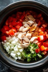 A top-down view of a slow cooker on a kitchen countertop filled with diced tomatoes, raw chicken pieces, chopped red bell peppers, and onions. The fresh and vibrant ingredients are coated with taco seasoning, ready to be mixed and slow-cooked to perfection.