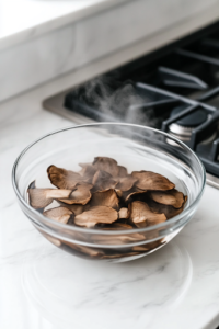 A glass bowl on a white marble cooktop filled with dried shiitake mushrooms soaking in steaming hot water. The mushrooms slowly expand as they absorb the liquid, becoming tender and rich in flavor.