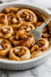 A close-up of the baking dish on a white marble cooktop, where warm caramelized apple filling is evenly spread over the flattened cinnamon roll layer. A spatula smooths out the mixture, ensuring every bite is packed with rich apple flavor.