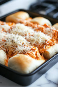 A close-up shot of a black baking dish on a white marble cooktop, where the bottom half of dinner rolls is neatly arranged. The prepared buffalo chicken mixture is evenly spread over the rolls, with a generous layer of shredded cheddar and pepper jack cheese sprinkled on top, ready for assembly.