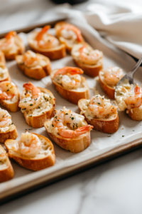 A baking sheet lined with parchment paper on the white marble cooktop, neatly arranged with baguette slices. A spoon is spreading the shrimp and cheese mixture evenly onto each slice, preparing them for baking.