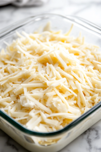 A close-up view of shredded sharp cheddar cheese being generously sprinkled over the assembled casserole in a glass baking dish on the white marble cooktop. The cheese is spread evenly, covering the surface, ready to melt into a golden, bubbly layer when baked.