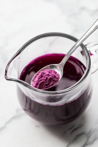 A close-up shot of a teaspoon of grape JELL-O powder being sprinkled into the glass pitcher on the white marble cooktop. The fine purple powder dissolves slowly into the liquid, deepening the color and adding a rich, fruity essence to the Potion Punch.