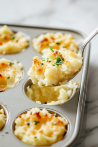 A close-up of a muffin tin on a white marble cooktop, with shredded Parmesan cheese generously sprinkled on top of each filled cup. The oven is open in the background, ready to bake the potato bites to golden perfection.