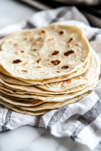 A close-up view of a stack of freshly cooked tortillas resting on a clean kitchen towel over the white marble cooktop. The tortillas remain warm and soft, ready to be served.