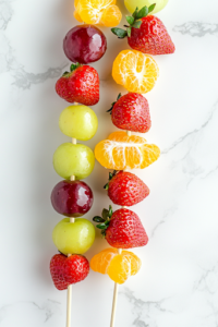 A close-up shot of five 12-inch wooden skewers lying on the white marble cooktop, each carefully threaded with alternating strawberries, grapes, and clementine segments. Small gaps are left between each fruit to allow for an even and smooth candy coating in the next steps.