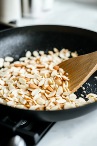 A close-up shot of a black skillet on a white marble cooktop, where slivered almonds are being toasted over medium heat. The almonds are turning lightly golden as a wooden spatula gently stirs them, ensuring even roasting.