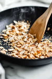 A black skillet on a white marble cooktop toasts crushed ramen noodles, slivered almonds, and sesame seeds. The mixture turns golden brown as steam rises, while a wooden spatula stirs gently for even roasting.