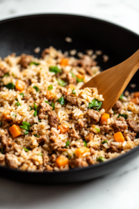 A close-up shot of the black skillet on the white marble cooktop, as uncooked long-grain rice is stirred into the meat and vegetable mixture. The grains begin to toast, turning a light golden brown, absorbing the rich flavors.