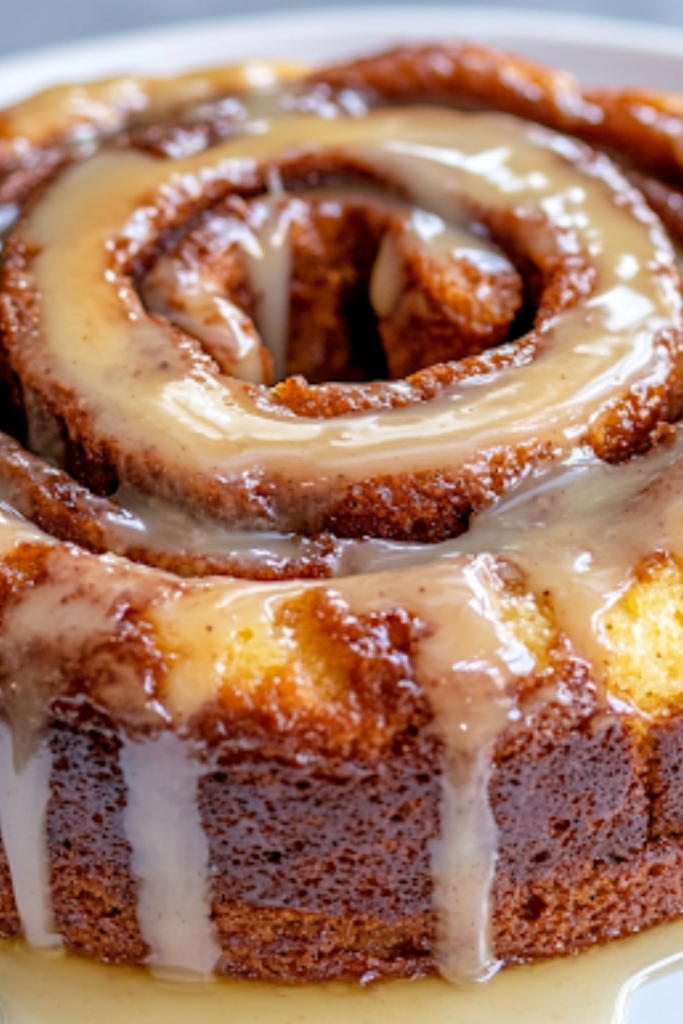 A close-up view of a warm cinnamon roll cake being drizzled with a rich vanilla glaze from a spoon. The glaze cascades down the cinnamon swirls, creating a glossy, sweet finish that enhances the cake’s soft and fluffy texture.