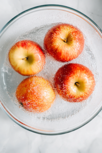 A close-up shot of four fresh medium apples being washed under running water over the white marble cooktop. A small knife and corer are placed beside them, ready for coring and preparation.