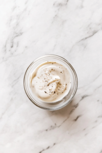 A small glass bowl on the white marble cooktop with mayonnaise, milk, sugar, salt, and pepper being whisked together. The dressing is smooth and creamy, ready to coat the salad ingredients.