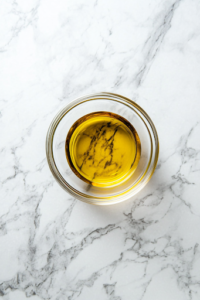 A close-up view of a small glass bowl on a white marble cooktop, where olive oil, red wine vinegar, Dijon mustard, garlic powder, onion powder, salt, and pepper are being whisked together into a smooth and flavorful dressing.