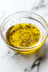 A close-up view of a small glass bowl on a white marble cooktop, where olive oil, lemon juice, dried oregano, and black pepper are being whisked together into a smooth and tangy dressing, ready to enhance the salad’s flavors.