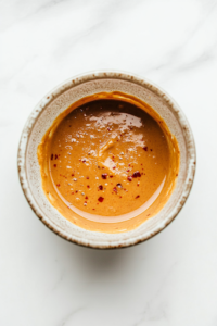 A close-up shot of a small mixing bowl on the white marble cooktop, with peanut butter, soy sauce, lime juice, water, sugar, and red pepper flakes whisked together. The ingredients blend into a thick, creamy peanut sauce, adding a rich and slightly spicy flavor.