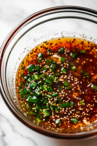 A close-up shot of a glass mixing bowl on a white marble cooktop, with tamari, toasted sesame oil, honey, grated ginger, chopped green onions, and sesame seeds being whisked together into a flavorful marinade for the Spicy Sesame Chicken Rice Bowl.