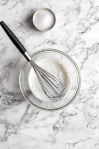 A small glass bowl on the white marble cooktop with white vinegar, sugar, salt, and black pepper being whisked together into a smooth and tangy dressing. A small whisk is in motion, blending the ingredients evenly.