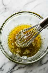 A close-up view of a glass bowl on a white marble cooktop as white sugar, vinegar, salt, and celery seed are whisked together. The smooth and glistening mixture is ready to be used as a sweet and tangy dressing for the salad.
