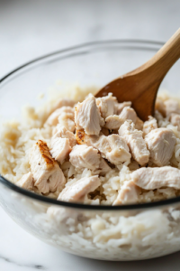 A glass mixing bowl on the white marble cooktop, now with bite-sized pieces of skinless, boneless chicken breast added to the creamy rice mixture. A wooden spoon stirs gently, evenly coating the chicken.