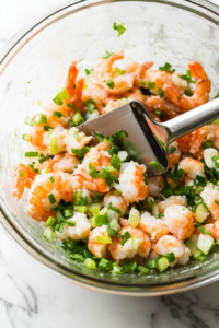 A close-up shot of the same glass mixing bowl on the white marble cooktop, now containing the drained crab meat, small shrimp, and freshly chopped green onions. A spatula is gently folding the ingredients together, ensuring even distribution.
