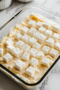 A close-up shot of a glass baking dish on the white marble cooktop, with small, evenly spaced cubes of cream cheese placed over the cake mix layer. The creamy white cubes stand out against the yellow cake mix.