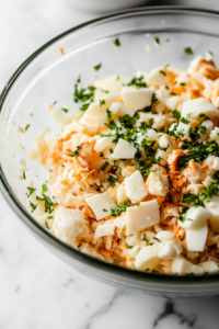Close-up of the same glass mixing bowl on the white marble cooktop, now with diced hard-boiled eggs and finely diced onion added on top of the crab meat. The ingredients look fresh and evenly cut.