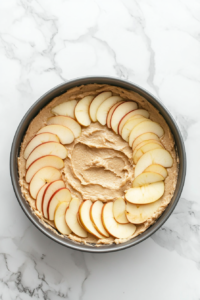 A close-up shot of a greased 9-inch round cake pan over the white marble cooktop, with cake batter spread evenly inside. Fresh Granny Smith apple slices are being carefully layered on top.
