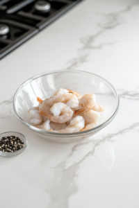 A close-up shot of raw peeled and deveined shrimp in a glass bowl over the white marble cooktop. A small dish of salt and black pepper sits beside it, ready for seasoning.