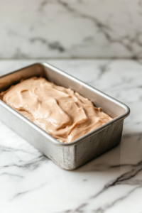 A greased loaf pan on a white marble cooktop, filled with smooth strawberry-rhubarb batter, ready to go into the oven.