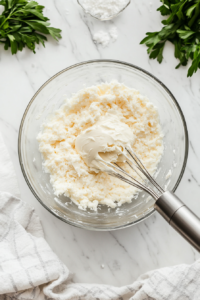 A glass mixing bowl on a white marble cooktop as softened butter and cream cheese are being beaten into a smooth, creamy mixture. A hand mixer is partially visible.