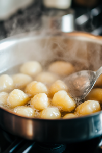 A separate pot on the white marble cooktop, filled with boiling water. Soft potato gnocchi are floating near the surface, just about to be drained with a slotted spoon. Steam rises from the pot, creating a cozy kitchen ambiance.