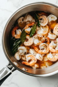 A close-up shot of a large stainless steel saucepan on the white marble cooktop, filled with shrimp shells, garlic salt, black pepper, bay leaves, and filtered water. The ingredients are floating as the water begins to warm.