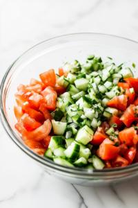 A clear glass mixing bowl placed over the white marble cooktop, filled with chopped tomatoes, diced cucumber, and finely chopped onion.