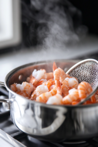 A close-up shot of a metal strainer draining the cooked shrimp over the stainless steel saucepan on the white marble cooktop. The shrimp are fully pink, and excess steam rises as they are lifted out of the broth.