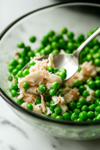 Close-up of a spoon gently folding bright green peas into the crab salad inside the glass mixing bowl on the white marble cooktop, ensuring they remain whole.