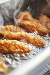 Close-up of battered fish fillets frying in bubbling hot oil inside a deep fryer on the white marble cooktop. The golden crust is forming as steam rises.